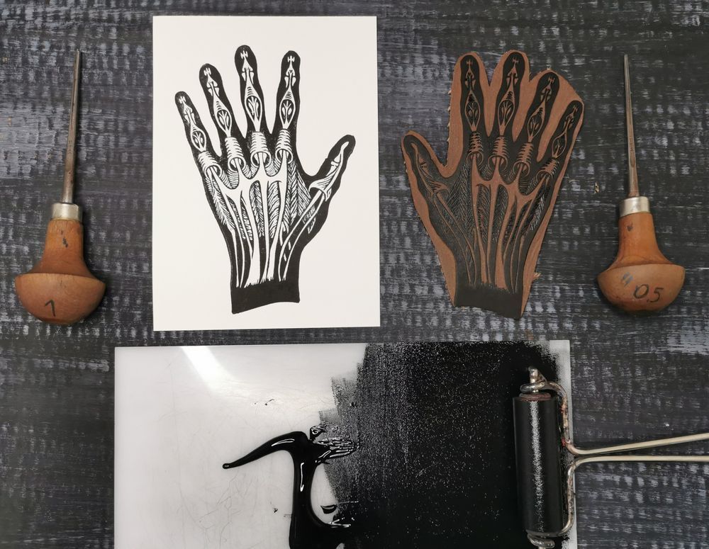 Auf dunklem Hintergrund liegt ein Linoldruck mit schwarzer Farbe auf weißem Papier. Das Motiv ist eine menschliche Hand. Da wo die Knochen, Sehnen, Muskeln wären, befindet sich keine Farbe und scheint somit als weiß durch. Rechts daneben liegt der dazugehörige Linolblock mit schwarzer Farbe versehen. Darunter liegt eine weiße Acrylplatte auf der schwarze Farbe ausgerollt wurde. Die verwendete Farbwalze liegt darauf. Links und rechts von der Anordnung liegen jeweils ein Linolschneidewerkzeug.