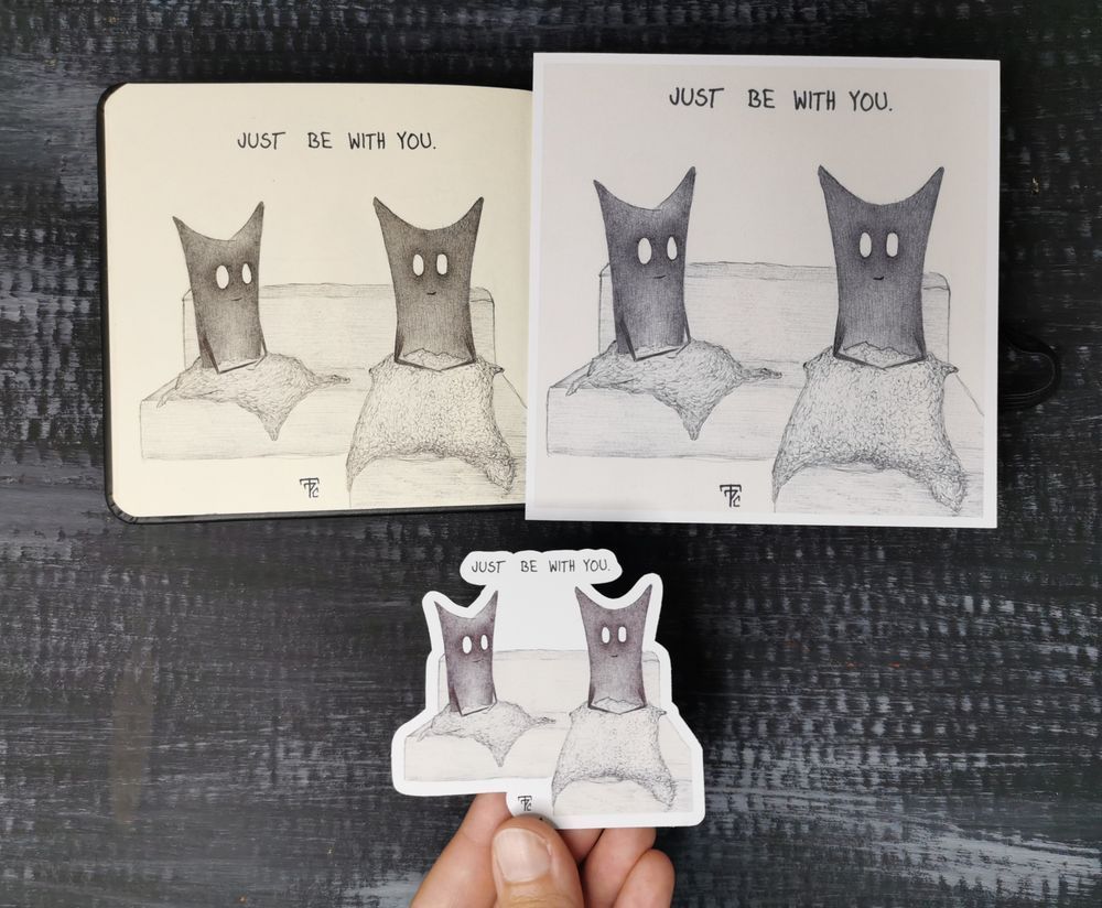 Auf dunklem Hintergrund liegt ein geöffnetes Skizzenbuch. Auf der linken Buchseite ist eine Zeichnung mit schwarzem Kugelschreiber. Das Motiv sind zwei geistartige Figuren mit großen, leeren Augen und zwei Spitzen am Kopf. Sie sitzen auf einer L-förmigen Couch. Die rechte Figur sitzt auf dem L-Stück. Beide haben eine Decke auf sich und schauen freundlich. Oben drüber steht folgender englischer Satz: just bei with you.
Auf der rechten Buchseite liegt ein Kunstdruck des gleichen Motives. Unter dem Skizzenbuch hält eine Hand einen Sticker, ebenfalls mit dem Motiv.
