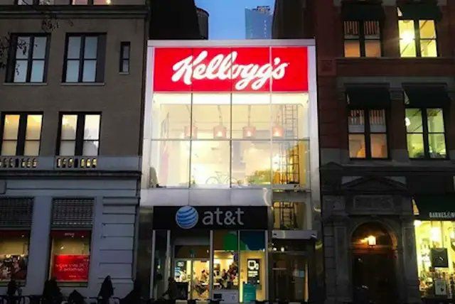 A bright, wall made of windows with the red and white Kellogg's logo above it, on a NYC street