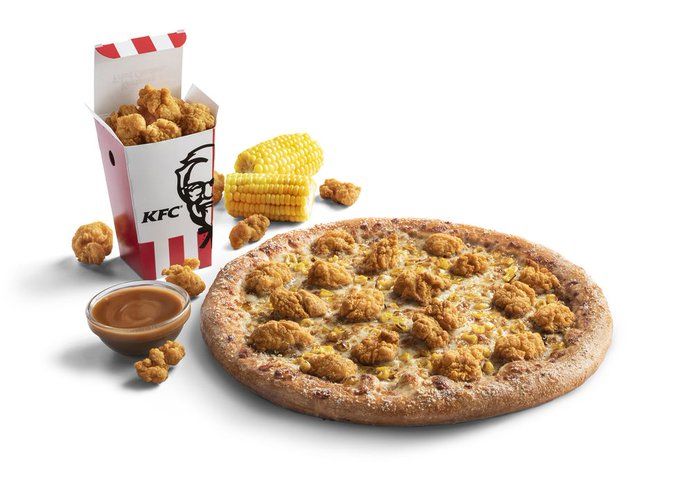 A round pizza topped with popcorn chicken, cheese, and corn. Next to it sits a bowl of gravy, a popcorn-style box with the KFC logo, filled with fried chicken, and an ear of corn