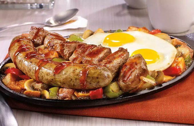 On a black skillet sits a large sausage covered in a thick brown glaze, next to a skewer of steak chunks and 2 over-easy eggs. on top of a bed of veggies