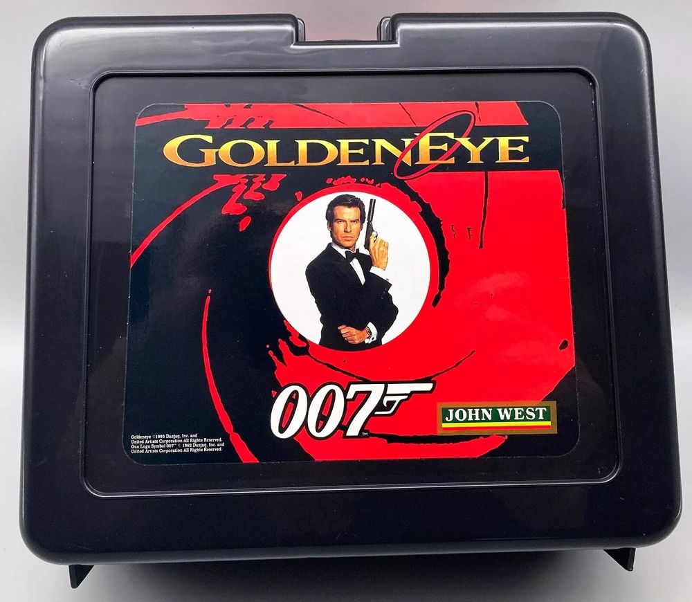 A black plastic lunchbox with the classic black/red "scope" James Bond intro logo, with Pierce Brosnan as James Bond, in the middle, holding a gun. At the top is the "GoldenEye" logo, with the 007 logo at the bottom