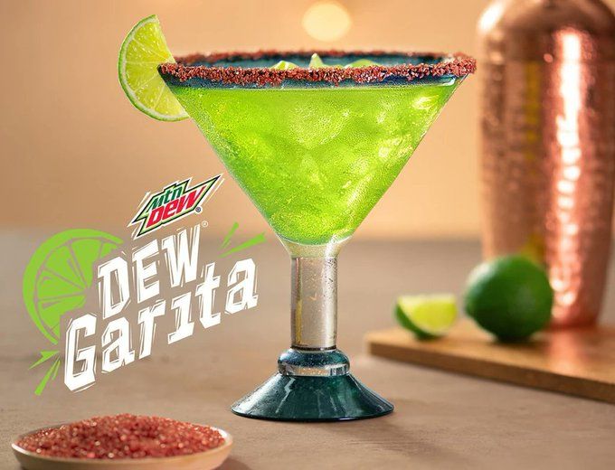 A stemware glass, filled with green liquid. There is a dusting of red sugar on the rim, and a slice of lime. Flanking the glass is a small dish of red sugar crystals, and a cutting board with a lime and drink shaker