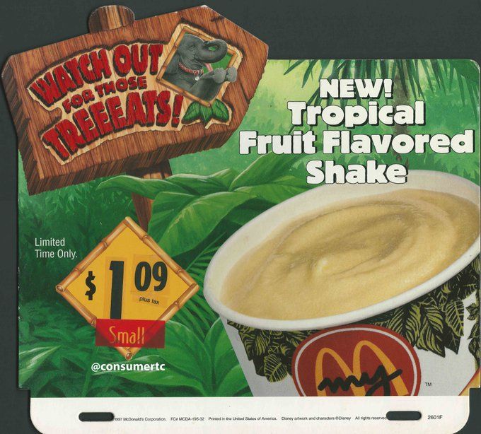 A McDonald's cup filled with a thick, creamy, light yellow substance, in front of a rainforest background. Above it is a wooden arrow sign pointing to the right, with the image of an elephant and the phrase "Watch out for those treeeats" carved into it