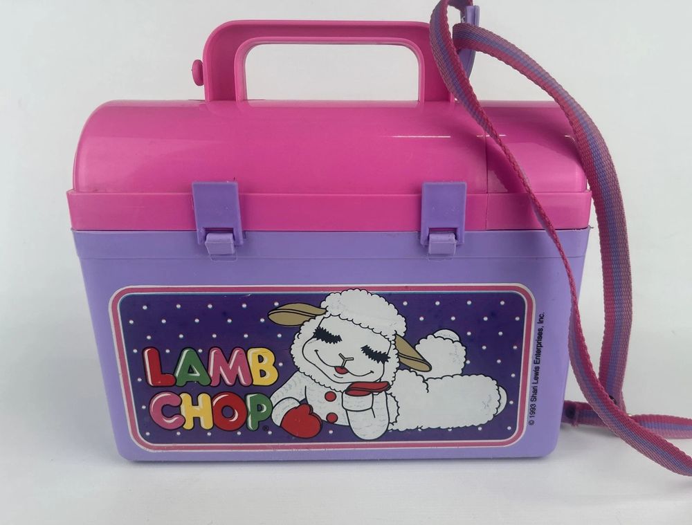 A purple and pink "worksite"-style lunch pail, with a purple and pink strap connected to the handle. On the body of the lunchbox are the words "Lamb Chop" written in brightly colored letters, against a blue and white dot background. An image of Lamb Chop, a white lamb puppet with long eyelashes and red mittens, reclines