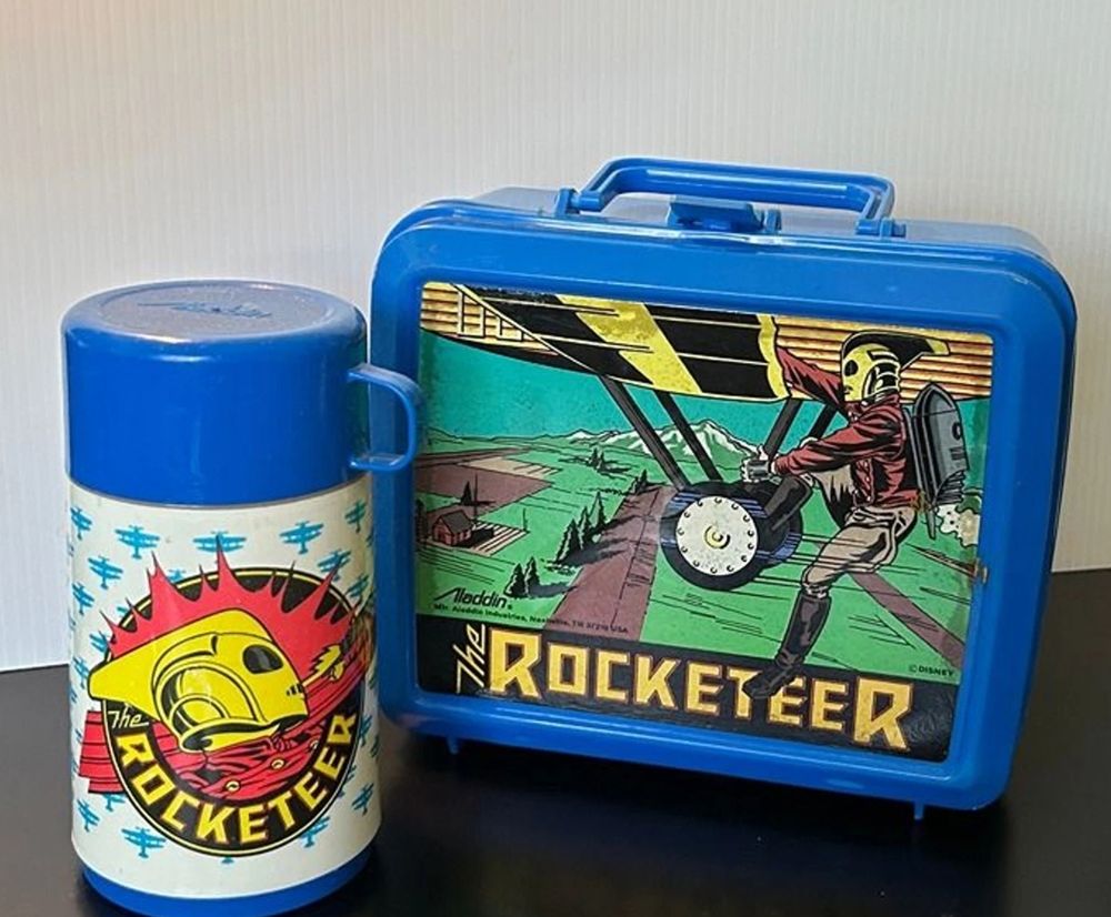 A blue plastic lunchbox with the illustration of The Rocketeer in his bomber jacket, boots, gold helmet and jet pack, hanging onto the wheels of a plane. Next to it is a white and blue thermos with an illustration of the Rocketeer's helmet