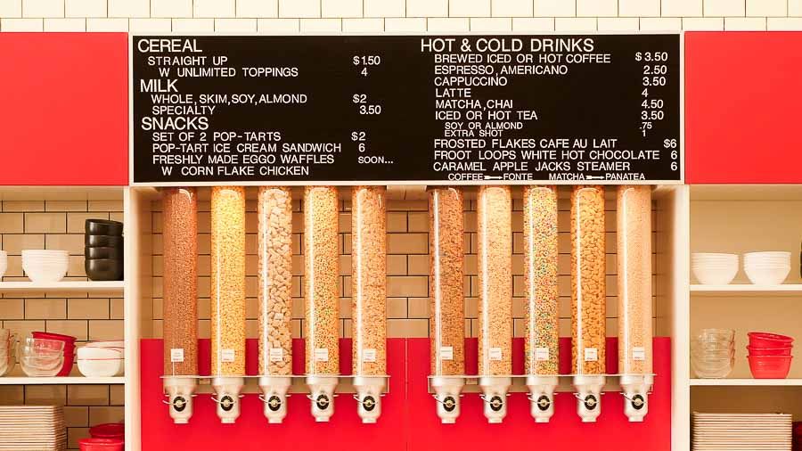 A black and white menu of cereal options and drinks, above a line of dispensers filled with cereals