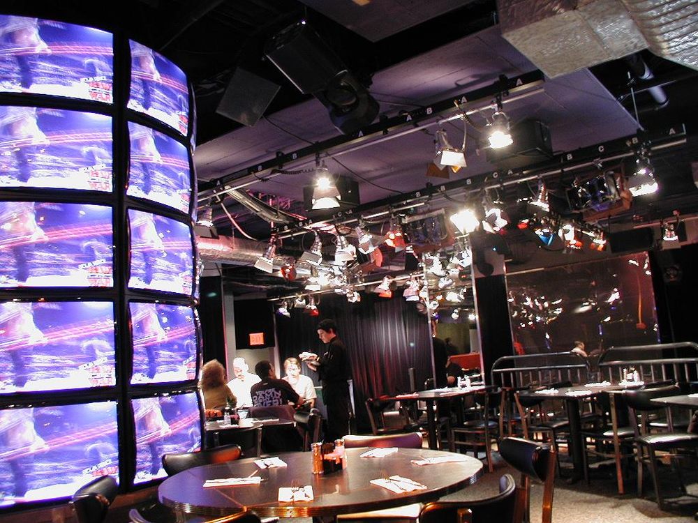 The inside of WWF New York, featuring round tables with chairs, stage-style light rigs, and a wall of TVs