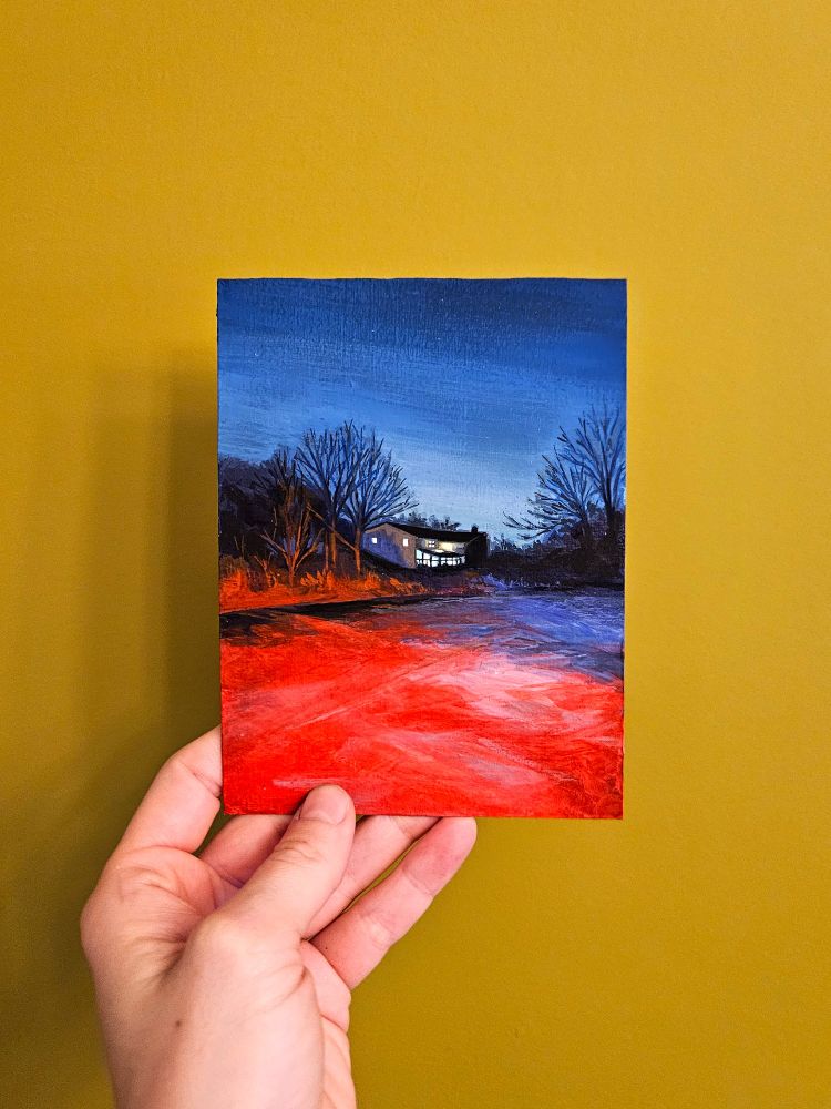 Image of a hand holding a small painting, made by Allegra Lockstadt, in front of a golden yellow wall. The painting is a night scene that shows a little house in the distance at dusk; the house has lights on and large trees stretch into a dark blue sky. The foreground is lit by the red glow of car tail lights, and in the distance, the colours switch to dusty blues. 