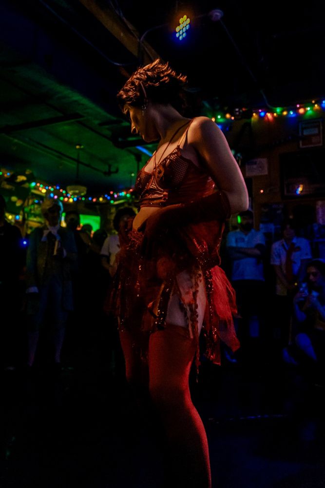 Colorful and dynamic shot of a burlesque-style dance performance by a local drag artist. The performer sparkles, scantily-clad in their stunning costume as they hold the attention of an enraptured audience. 