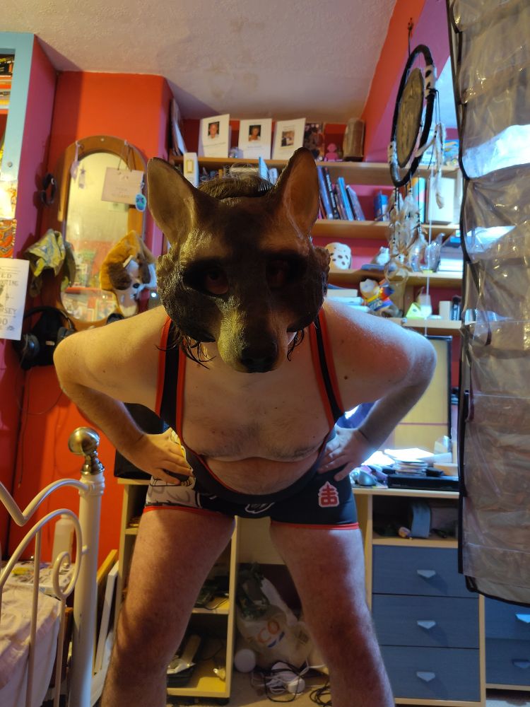 Man in raccoon mask wearing a singlet and leaning over directly at the viewer