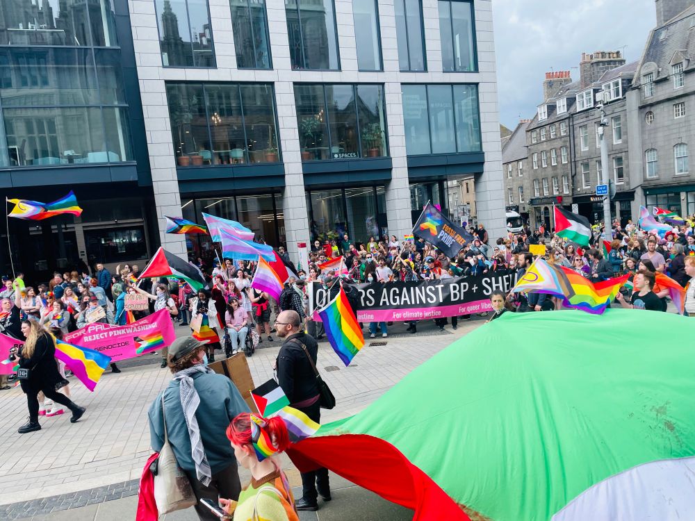 A large crowd march past Marischal Square holding many Pride, trans and Palestinian flags. A banner saying - “Queers against BP + Shell” - can be seen.