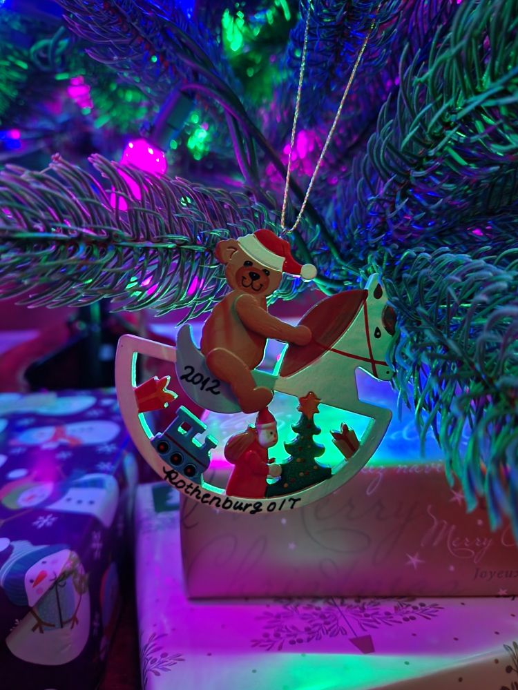 Christmas Ornament of a wooden rocking horse that is white with a red mane. Sitting on the horse is a brown teddy bear wearing a red and white santa hat. Below it are some toys and santa.