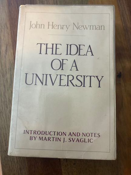 cream-colored book cover set off against the darker background of the wooden desk upon which it rests. the book is John Henry Newman's The Idea of a University, with introduction and notes by Martin J. Svaglic. a classic.