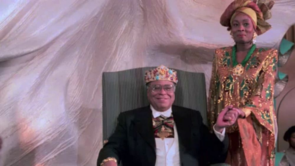 a man wearing a crown is sitting in a chair next to a woman in a colorful dress