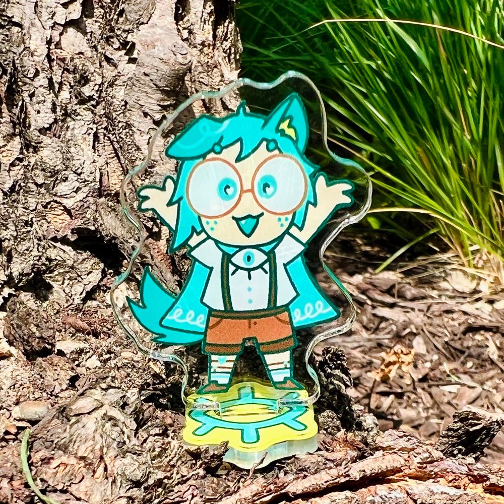 Photo of an acrylic stand collectible. The art features Teal Emile, a gender-ambiguous character with blue hair and puppy features. They are wearing glasses, suspenders, shorts, and a cape. They are raising their hands excitedly into the air.

The photo was taken in front of a tree and various plants.