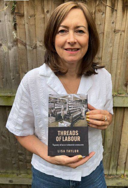 DePOT affiliate Lisa Taylor, stands in front of a wooden fence similing, wearing a white blouse and blue jeans, holding a copy of her new book: "Threads of Labour: Tapestry of an Ex-Industrial Community"