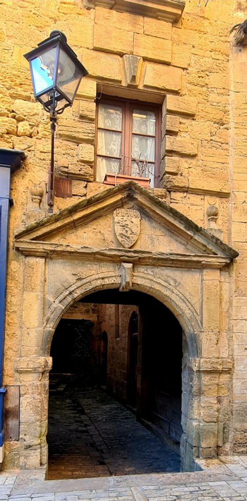 C’est un vieux passage voûté en pierre, typique d’une ville médiévale, en l'occurrence Sarlat. 
Au-dessus de l’arche, on voit un fronton triangulaire avec un blason sculpté.
Une fenêtre ancienne avec des volets en bois se trouve juste au-dessus, et une lanterne en fer forgé est fixée sur le côté.
Le passage donne sur une petite ruelle sombre pavée en pierre.