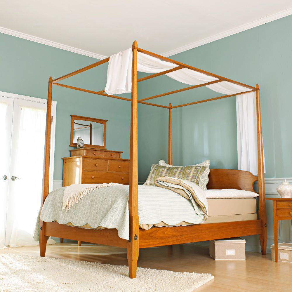 Elegant Shaker-style pencil-post bed handcrafted from warm-toned wood, shown in a serene bedroom with soft green walls. The bed features slender tapered posts connected by canopy rails with draped white fabric. A matching wooden dresser and nightstand complete the minimalist, traditional aesthetic.