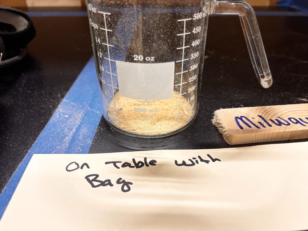 A detail shot of a testing procedure for a cordless mitersaw showing how much sawdust was left behind after cutting when using the saw's dust bag. The label "Milwaukee" is marked on a scrap of wood and a placard reading "On Table With Bag" sits next to a measuring cup holding the debris measuring how much dust remained on the worksurface after the cut.