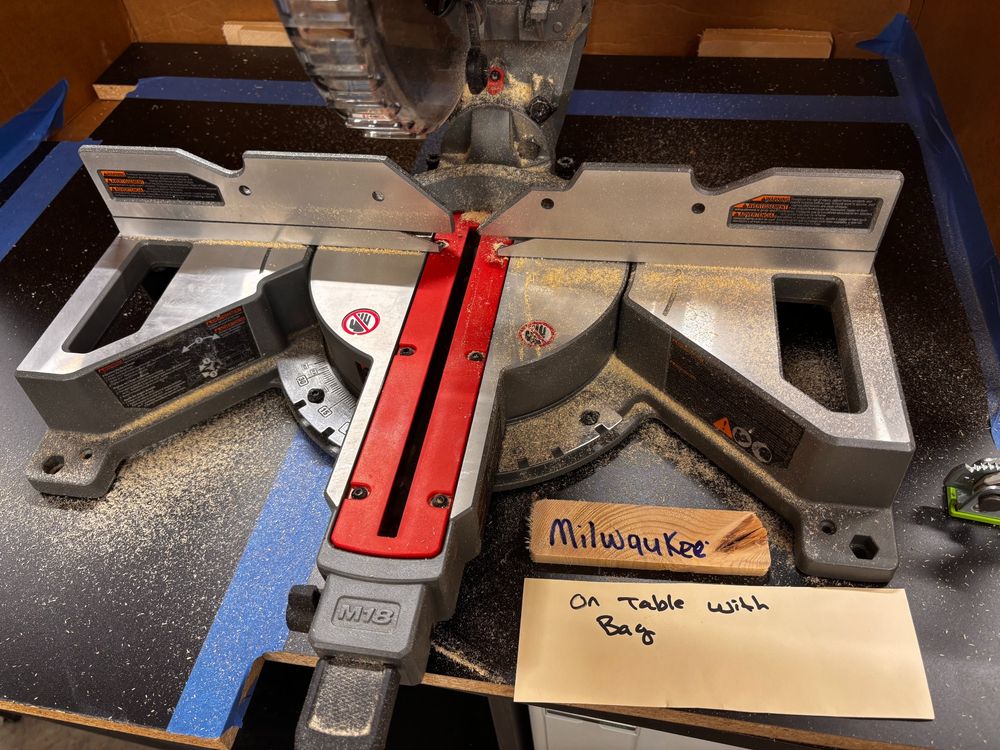 A detail shot of a testing procedure for a cordless mitersaw showing how much sawdust was left behind after cutting when using the saw's dust bag. The label "Milwaukee" is marked on a scrap of wood and a placard reading "On Table With Bag" sits next to the saw denoting how much sawdust remained on the worksurface after the cut.