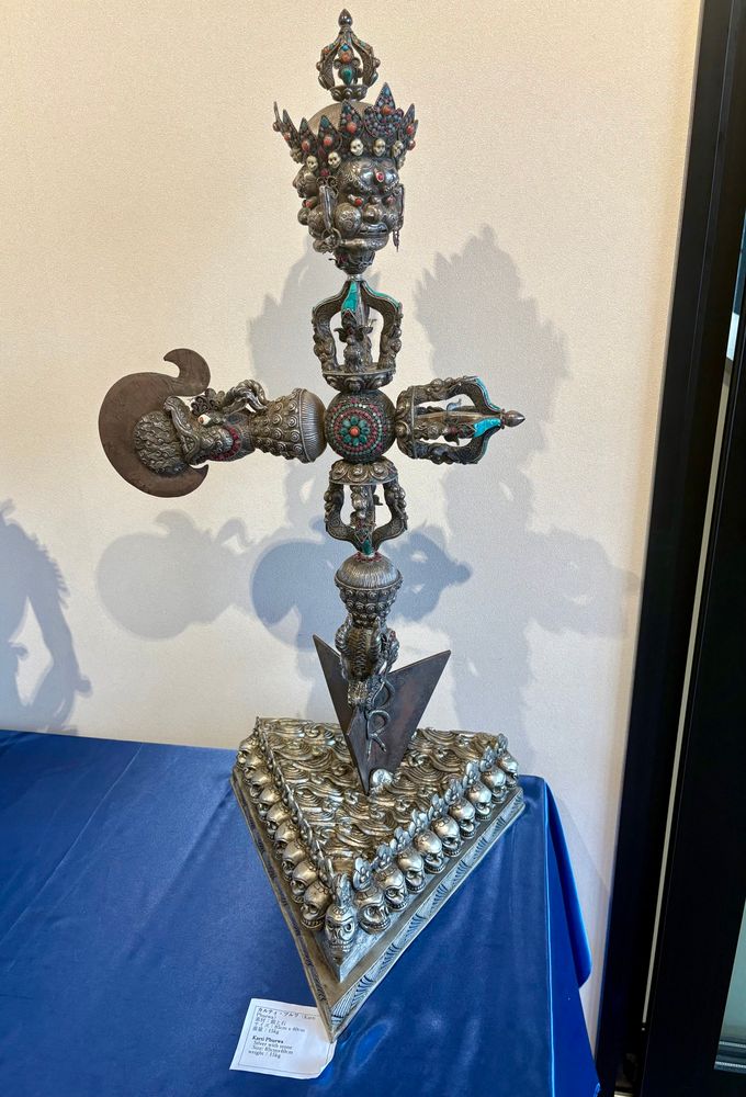 ネパール館に展示されていた、カルティ・ブルワという、おそらく仏教芸術の一種かと思われるが、詳細不明。銀と石製。髑髏が敷き詰められた三角形の台のほぼ中央に、鏃型の物が突き刺さる。突き刺さるもの全体を矢とするならば、矢羽の部分には髑髏の冠を被る多面の憤怒像。また、シャフトの中間部分には、左側が龍頭の三鈷杵のようなものが組み合わさっている。所々におそらくルビーと翡翠が嵌め込まれている。