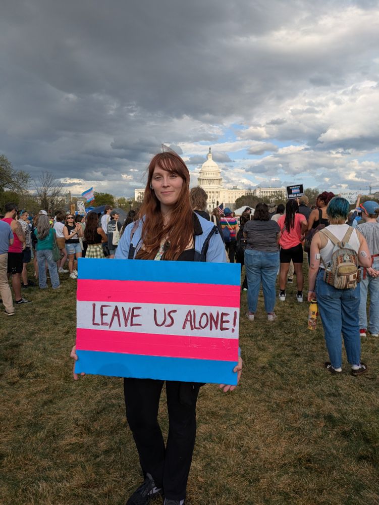 Myself, RunaMorgen holding my "Leave us Alone!" Transgender flag sign in front of the US capitol building on trans day of visibility, 2025. The sun shines over the trans rally, as the dark clouds collect over the capitol.