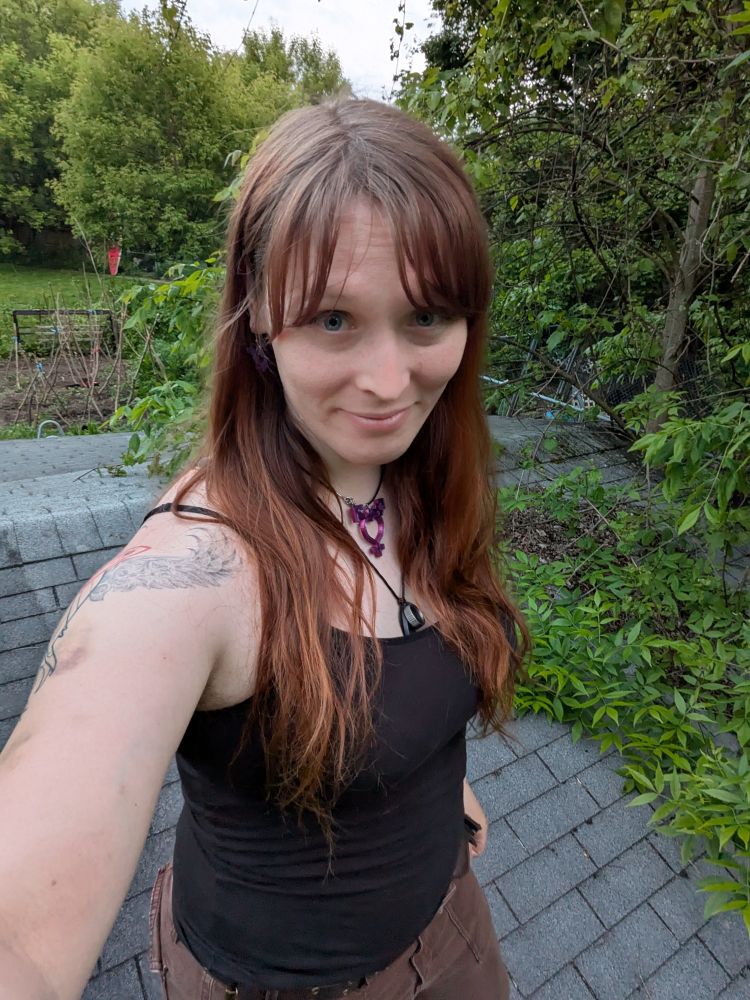 A selfie of me, a trans lady standing on an old roof in front of my garden, worn but happy, simple black tank top with holes, brown jeans, trans symbol necklace, long red-brown hair, covered in bruises from having trans girl skin.