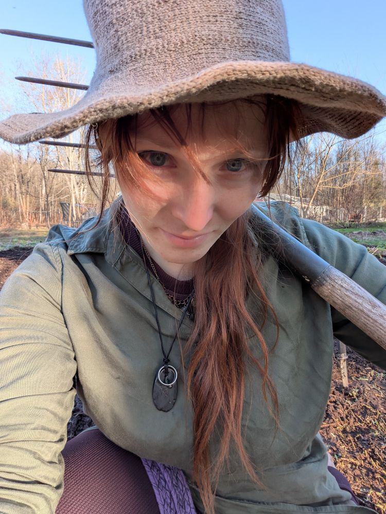 Me, a transgender woman, long brown-red hair in dirty green tunic & grey hat holding a pitchfork in front of a newly started garden in the late afternoon.