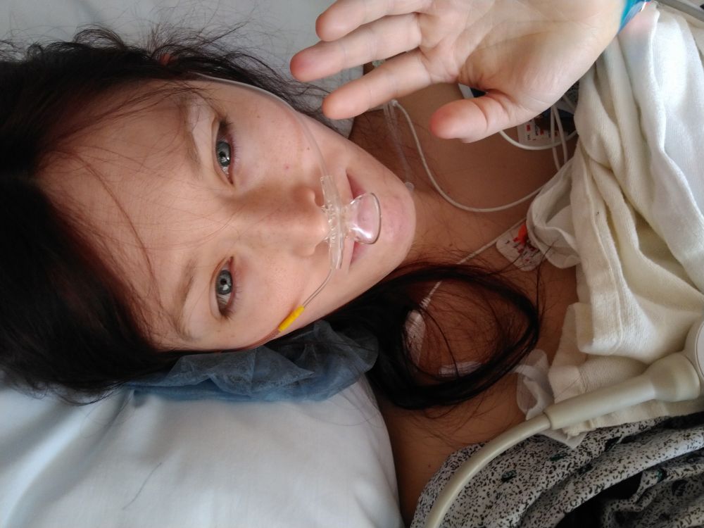 Myself, 4yrs younger, with breathing tube, long dark red hair, and hospital gown, lying on a hospital bed, with my hand up in a hello gesture, at peace and in my power for the first time after genital affirmation surgery.