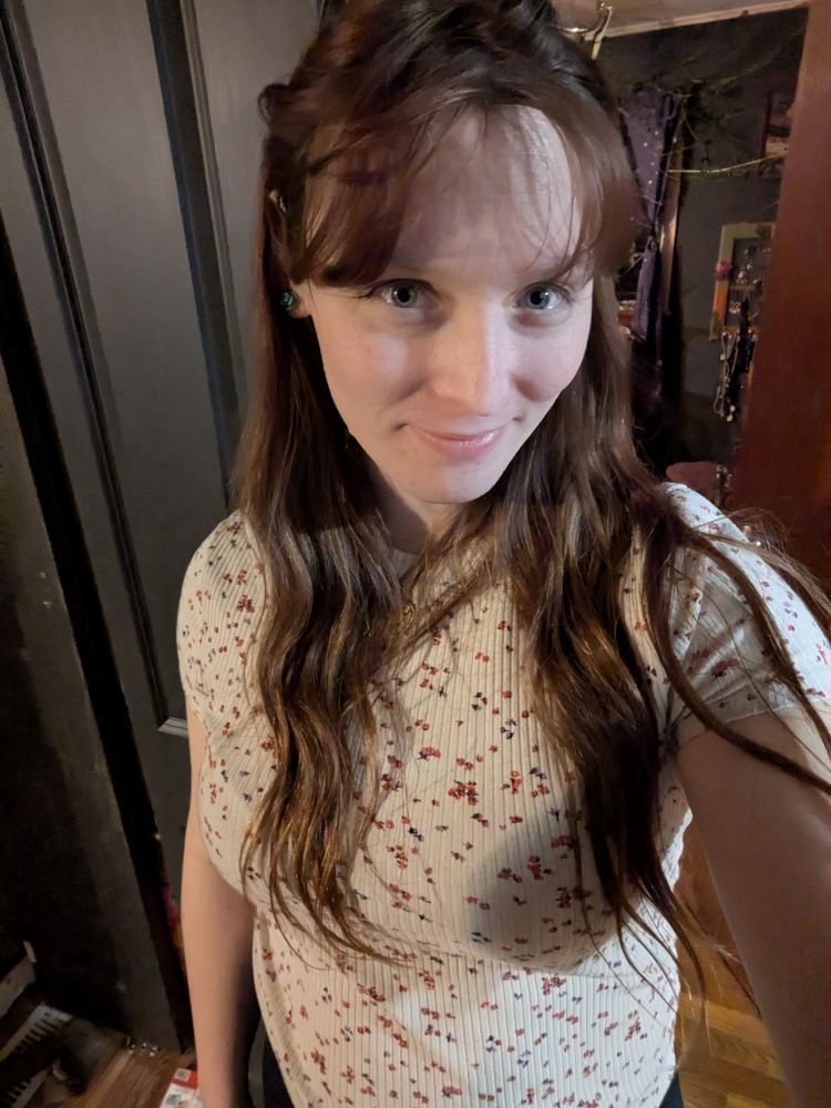 Me, a transgender woman with long brown hair, wearing a simple white flower blouse smiling after just catching a glimpse of my reflection & actually liking what i saw. 