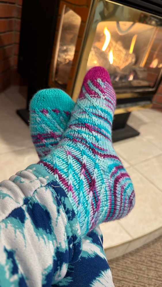 Feet wearing blue and purple knitted socks with spiralling slipped-stitch texture in front of a small gas fireplace.