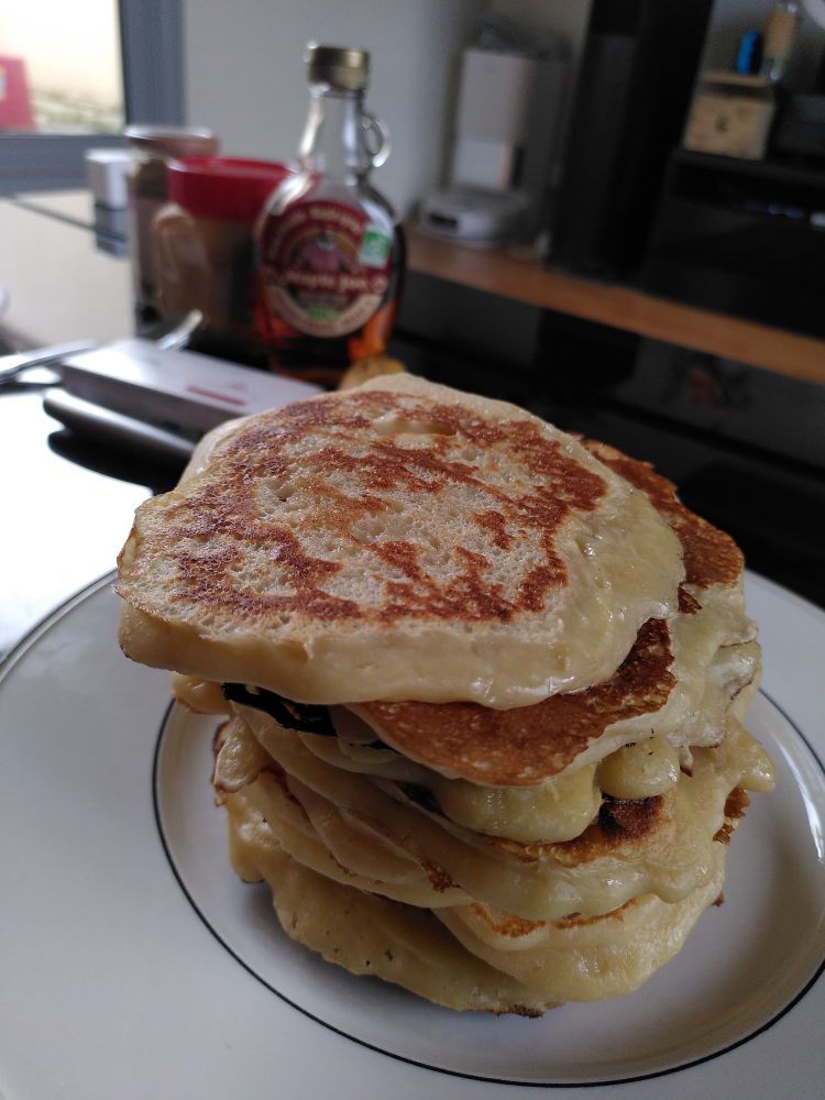 Une photo d'un tas de pancakes très appétissants, en fond on voit une bouteille de sirop d'érable et de beurre de cacahuète 