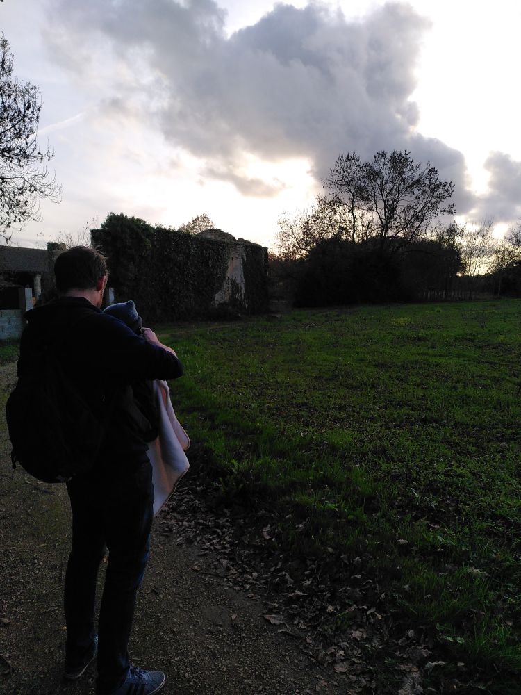 Photo d'un homme de dos, qui tient un bébé qu'on ne devine qu'au bonnet qui dépasse. En arrière plan, un champ et une vieille maison. La photo est prise au crépuscule.