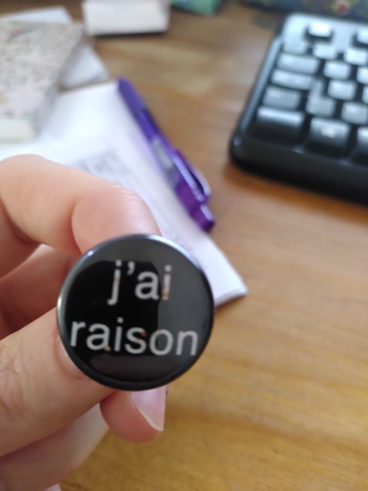 Photo en gros plan d'une main qui tient un badge rond et noir sur lequel est marqué "j'ai raison". Dans le fond on voit un bureau en bois sur lequel il y un clavier d'ordinateur, des papiers et un stylo.