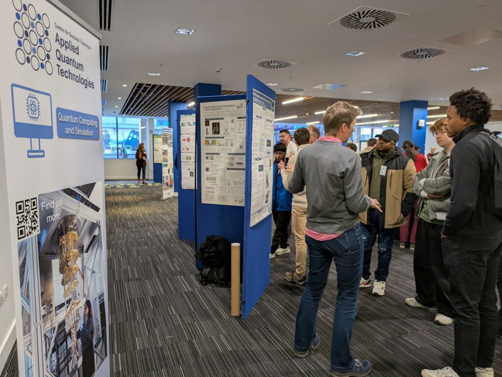 Banner on left for AQT cdt and people talking around poster boards in the centre and queuing for lunch on the right.