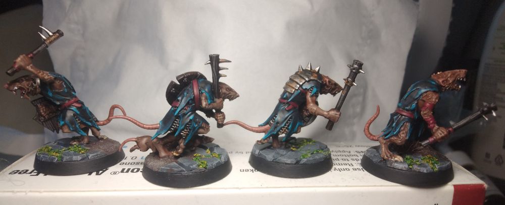 Four skaven clanrats wielding clubs and shields photographed in a row. The tiny ratmen are painted with brown fur, rusty worn metal armour, red belts, and tattered clothes layered with turquoise and bright blue highlights at the edges. They stand on mossy cobblestone and muddy bases.