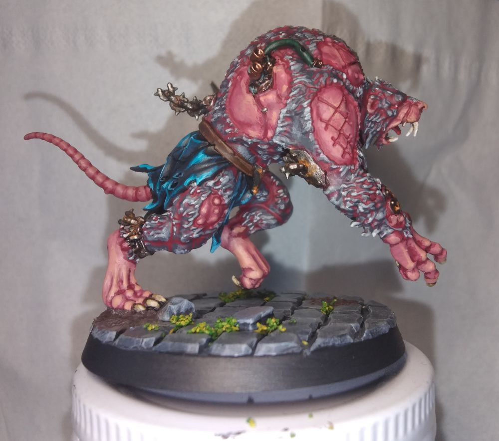Depicts the right-hand side of a giant mutated rat-man: a skaven rat ogor. It is in a running pose, toothy jaw open, right arm forwards with claws bared with a brass metal tube embedded into the flesh, left arm not visible. That left shoulder is covered in overlapping armour plates, similarly brown and battered and rusted. It is covered in carefully painted grey fur, with red in the shadows. Its fur splits apart, here visible on the upper arm, the back, and the thigh of the closest leg. The exposed skin beneath is horribly red and raw, with careful blends going from red to a pale peachy skin tone. Embedded in the back are some green tubes, running out from brass exhaust pipes also embedded. At it's waist there is a dark ragged cloth, starting black, with edges highlighted in progressively brighter teal tones. It's leg is shackled, with torn chain- torn chain emerges from plates grafted to its back and arm too. Both are rusted and dirty.

The base he stands on is mossy and muddy cobblestones.