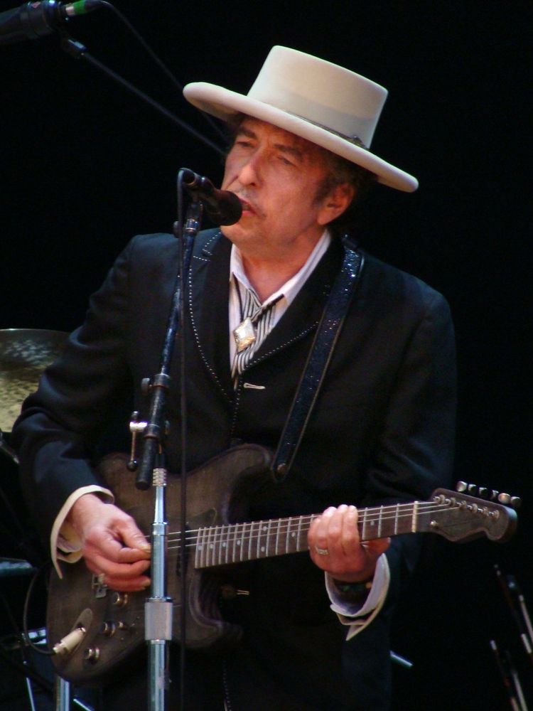 Bob Dylan auf der Bühne mit Gitarre und Mikrophon. Er trägt einen cremefarbenen Hut mit breiter Krempe