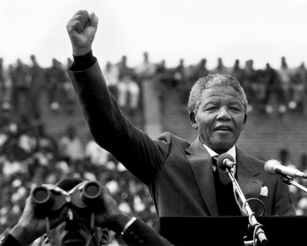 Nelson Mandela speaking to a crowd at a podium with his right fist in the air.