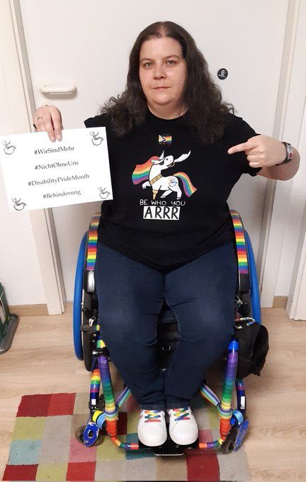 Ich sitze in meinem quietsch bunten Rollstuhl, trage ein T-Shirt mit einem Einhorn mit Regenbogenschweif und Mähne, es hat eine Augenklappe, einen Säbel im Maul und ein Holzbein.
Es lächelt.
Text auf Englisch.
Be who you arrrr.
In der Hand halte ich einen Zettel.
Text.
#WirSindMehr
#NichtOhneUns
#DisabilityPrideMonth
#Behinderung