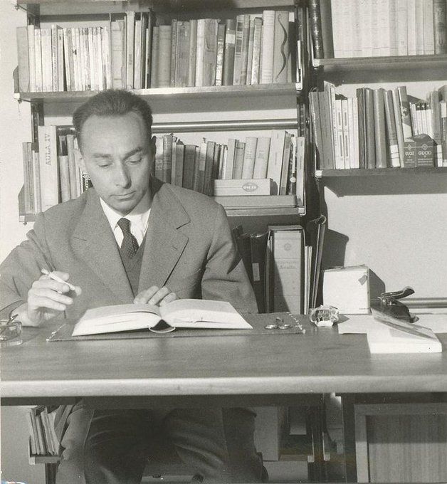 Primo Levi schreibend an einem Tisch, hinter ihm ein Bücherregal.