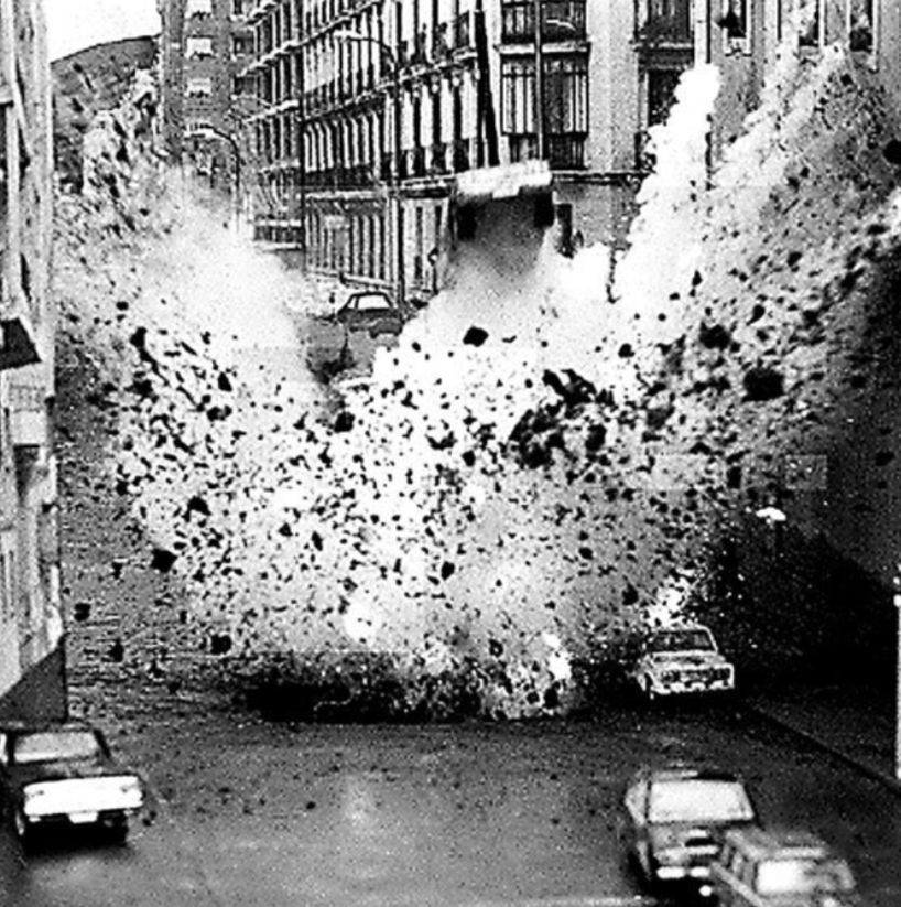 Grobkörnige Schwarz-Weiß-Fotografie. Eine Straßenschlucht, mehrgeschossige Häuser, am Rand parken Autos. In der Bildmitte findet eine gewaltige Detonation statt, die das Auto von Carrero Blanco etliche Meter in die Luft schleudert.