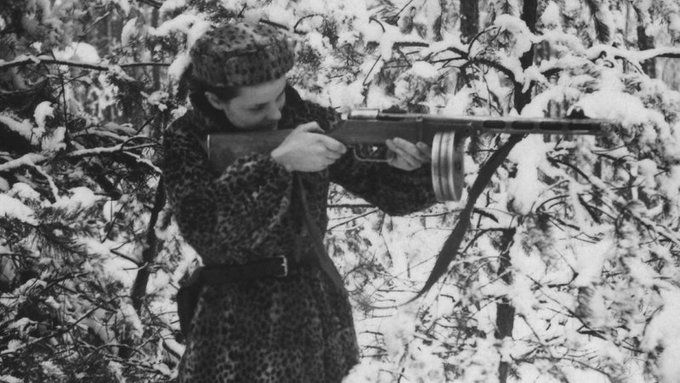 Fotografie, eine Frau mit Pelzmantel und -mütze zielt mit einer Maschinenpistole