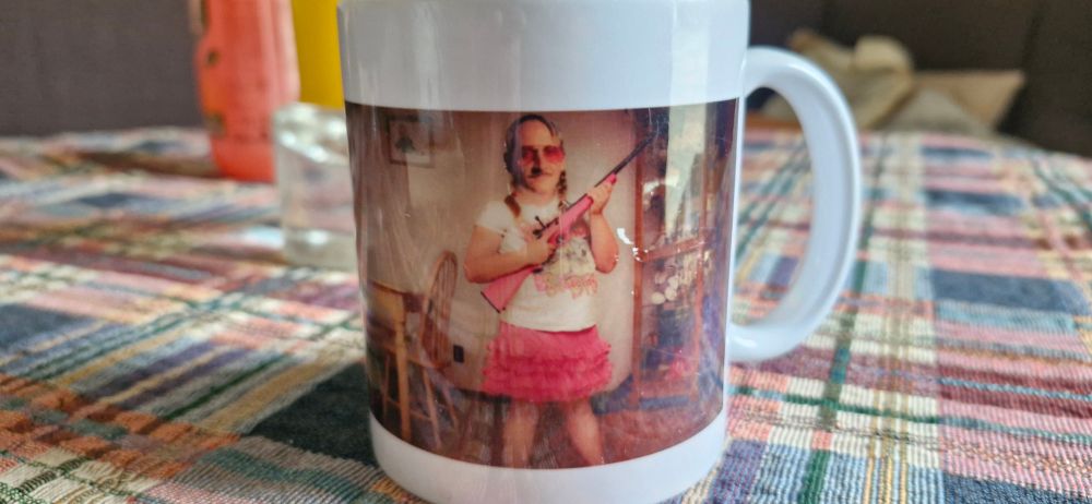 Tasse. Ein Mann mit Bart, Brille und Zöpfen trägt ein rosa Gewehr und einen dazu passenden Rock. 