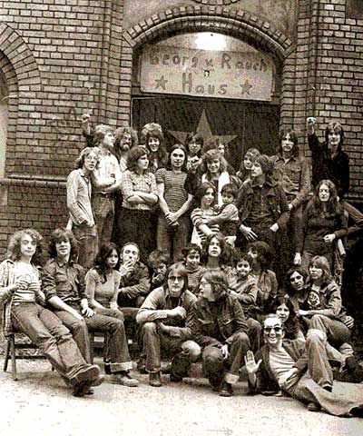 Schwarz-Weiß-Fotografie, Gruppenbild von etwa 30 Personen, viele mit langen Haaren, einige mit Kindern, vor einer Ziegelwand und einem Hauseingang, über dem steht: Georg v. Rauch-Haus.