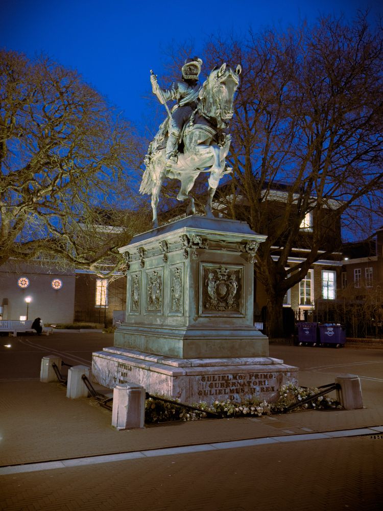 A nighttime photograph of an equestrian statue depicting a person riding a horse, located in a public square. The statue is placed on a large stone pedestal with inscriptions. Surrounding the statue are paved pathways and bare trees, indicating a season of late fall or winter. The sky is clear, with deep blue hues contrasting against the lit statue, highlighting its features and the surrounding architecture in the background.
