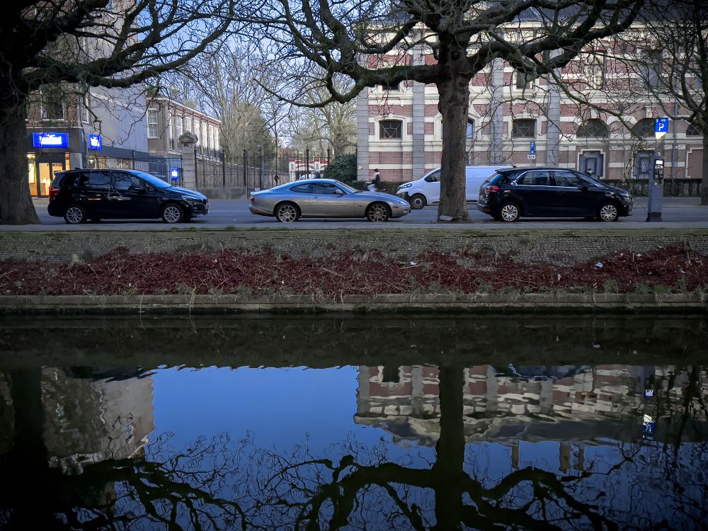 A street view featuring parked cars along the roadside, with leafless tree branches overhead. Across the street are several apartment buildings. In the foreground, a calm canal reflects the trees and buildings, creating a mirror-like effect.