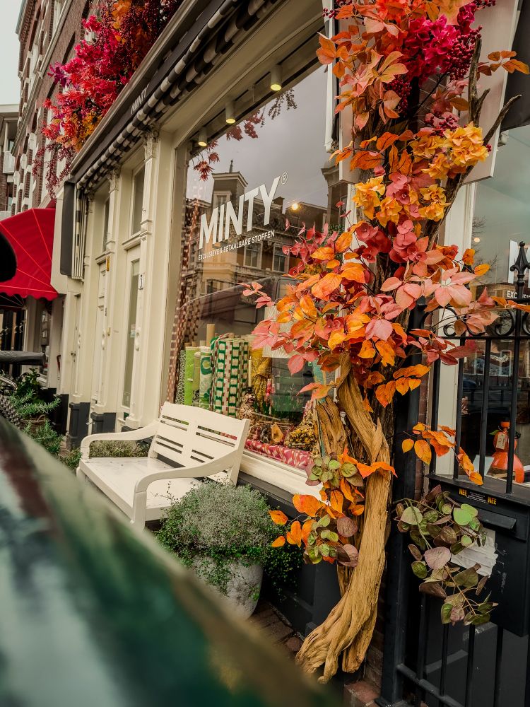 A charming street view featuring a cozy café with a vibrant autumn-themed floral display adorning its entrance. The café has large windows showcasing its inviting interior, and the sidewalk is embellished with planters. A bright red awning provides a splash of color, adding to the warm and welcoming atmosphere of the scene.