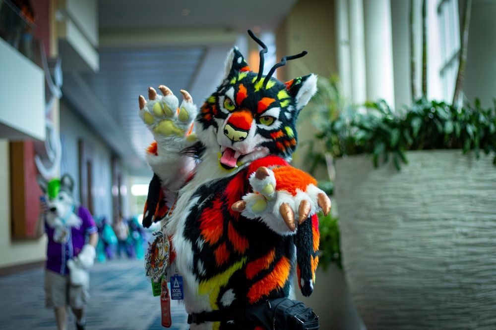 Enzo M. Tiger fursuiter, with a long hallway behind him at Megaplex 2025, standing next to a huge planter holding a tree, one paw raised behind and the other pointed at the camera